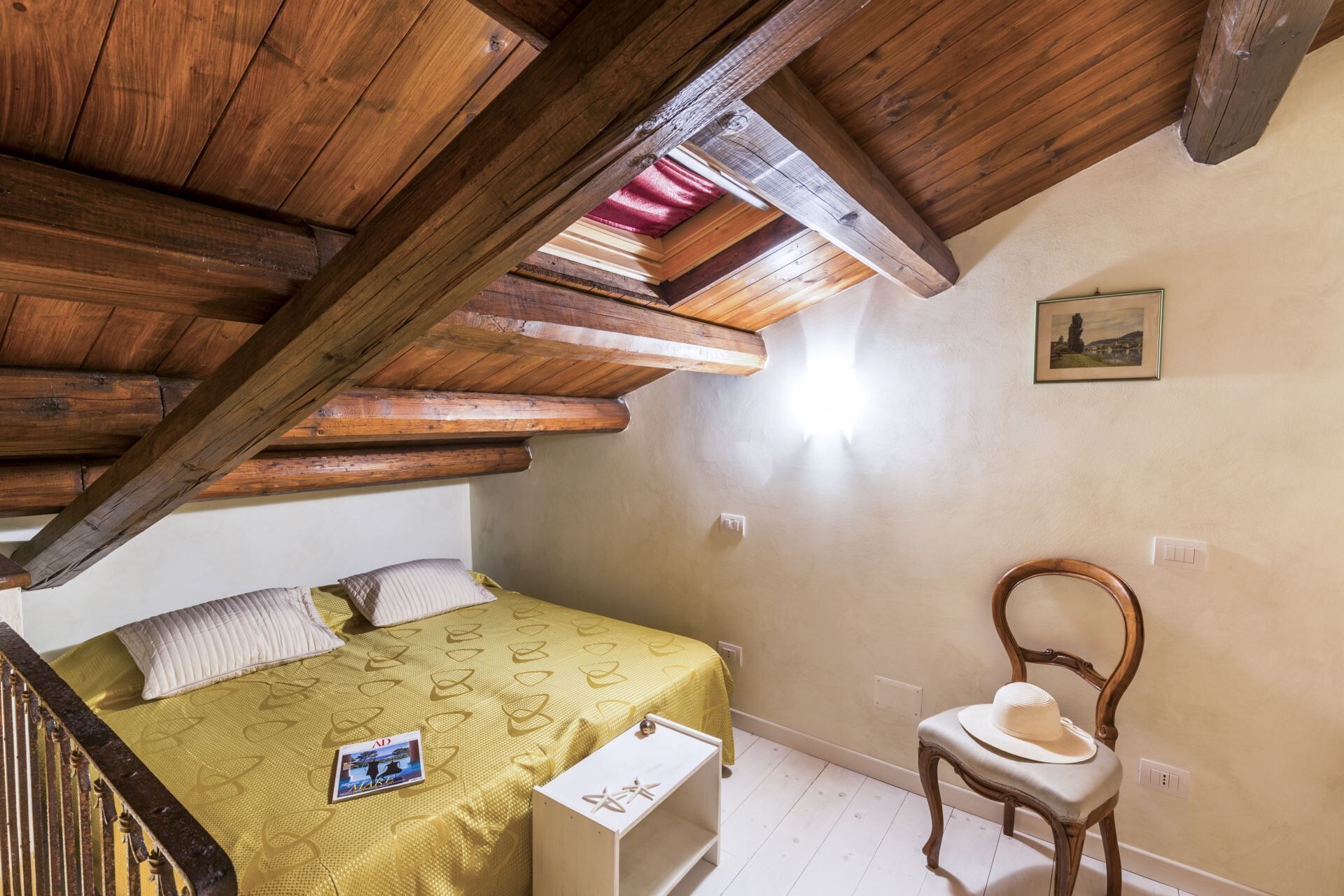 A bedroom with a sloped ceiling, a bed with a yellow patterned bedspread, a wooden chair, and a small table with a book and a box.