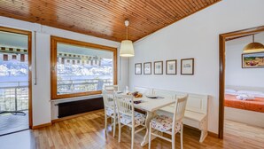 Un comedor con techo de madera, paredes blancas y un ventanal grande. Se observan sillas blancas, una mesa y una lámpara colgante.