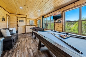 A wooden cabin interior with a pool table, a sofa, and large windows offering a view of the outdoors. - Luxe Cabin w/Privacy! Hot Tub* Fireplace + Firepit (Sevierville)