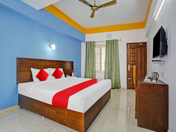 Habitación de hotel con cama de madera, ropa de cama roja y blanca, ventilador de techo, televisor y pequeña mesa de madera con tetera.