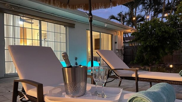 En solig utomhusavslappningsplats med en flätad loungestol, en metallhink, två glas och en hoprullad handduk på ett brickbord.