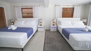 Ett hotellrum med två sängar, var och en med en vit sänggavel, ett blått överkast och en vit kudde. Det finns två sängbord med lampor, en grå matta och ett fönster med träpersienner.