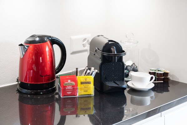 Una encimera de cocina con un hervidor eléctrico rojo, una cafetera negra, una jarra de vidrio, una taza y platillo blancos, y dos cajas de té Twinings.