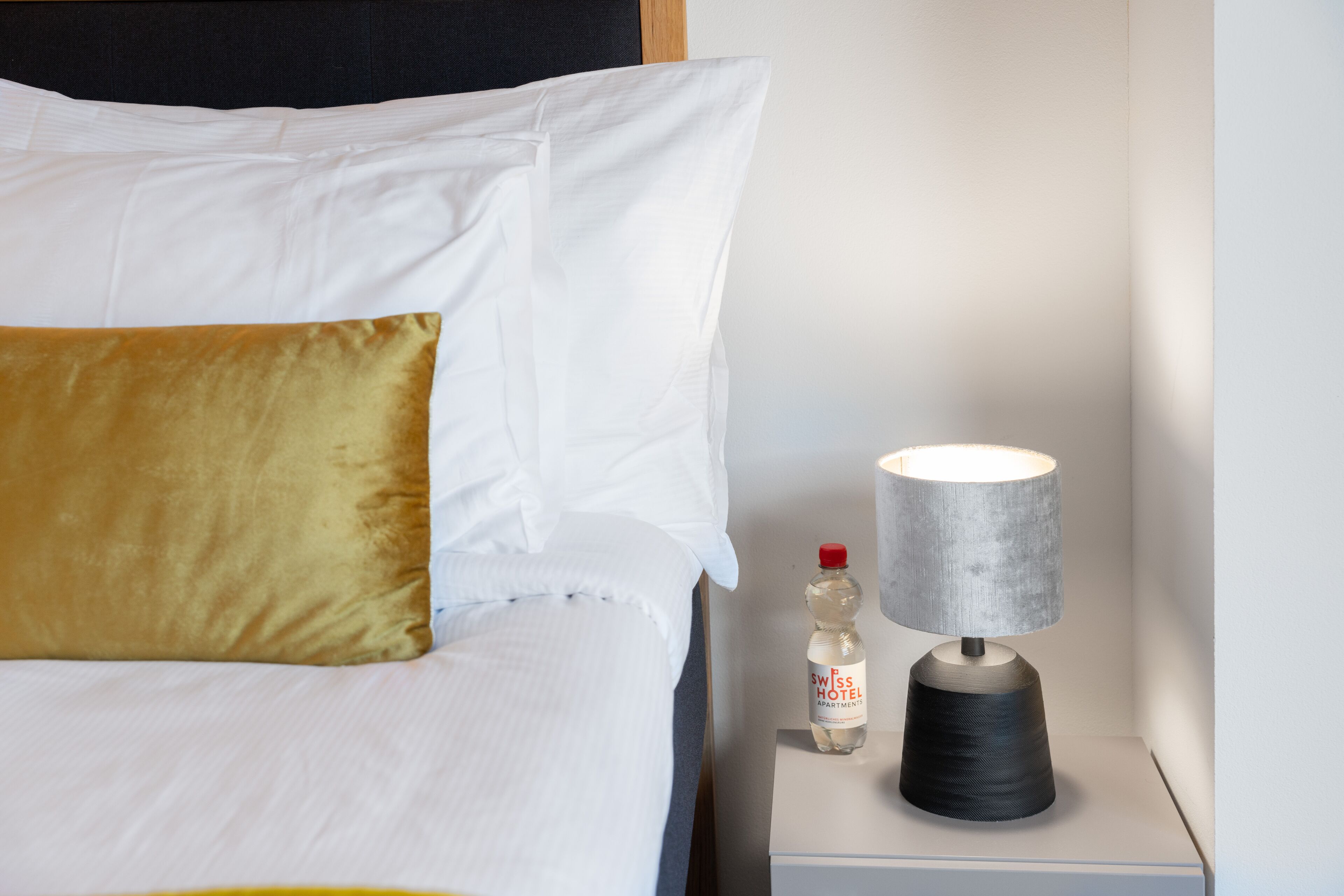 A neatly made bed with white and gold pillows, a bottle of water, and a lamp on a bedside table.