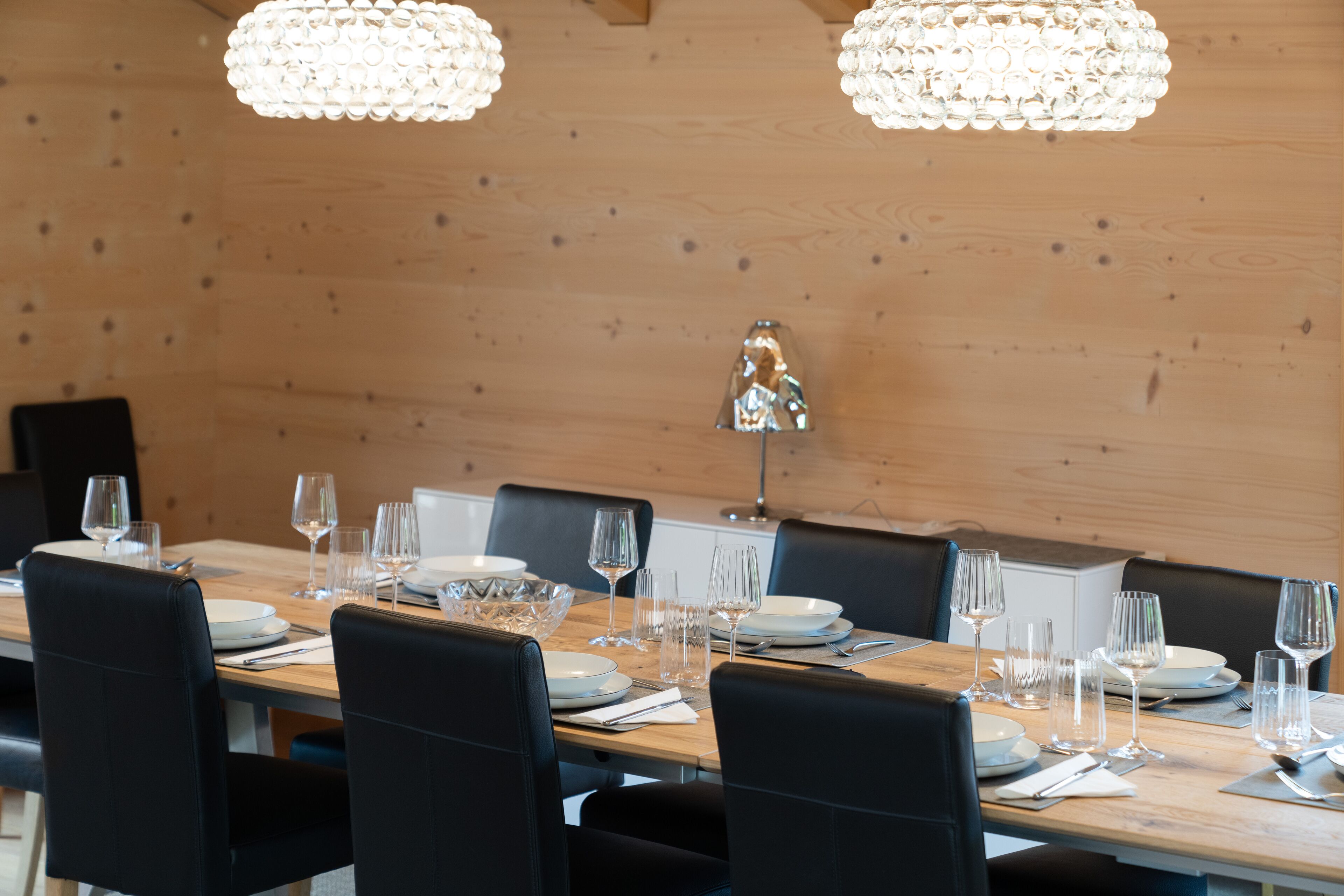 A dining area with a wooden table set for a meal, black chairs, and two crystal chandeliers.