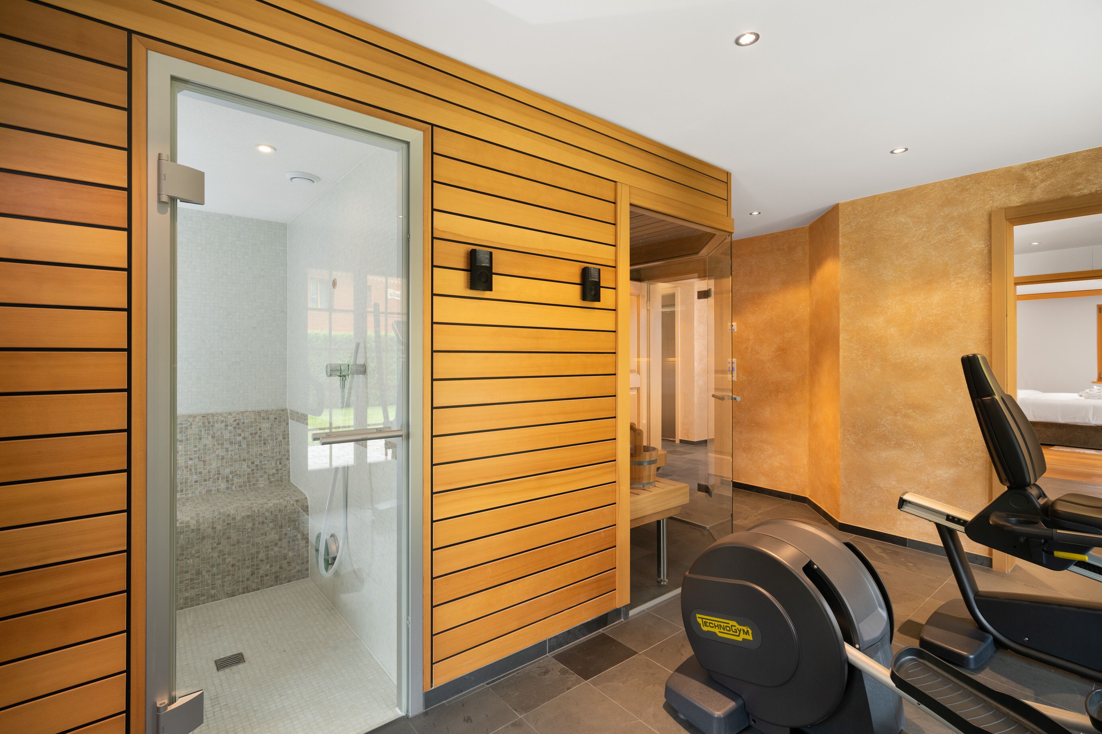 A modern hotel room with a wooden panel wall, a glass shower enclosure, a treadmill, and a bed with a view.