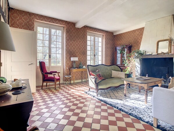 A living room with a checkered floor, a red armchair, a sofa, a fireplace, and a window with a view of trees.