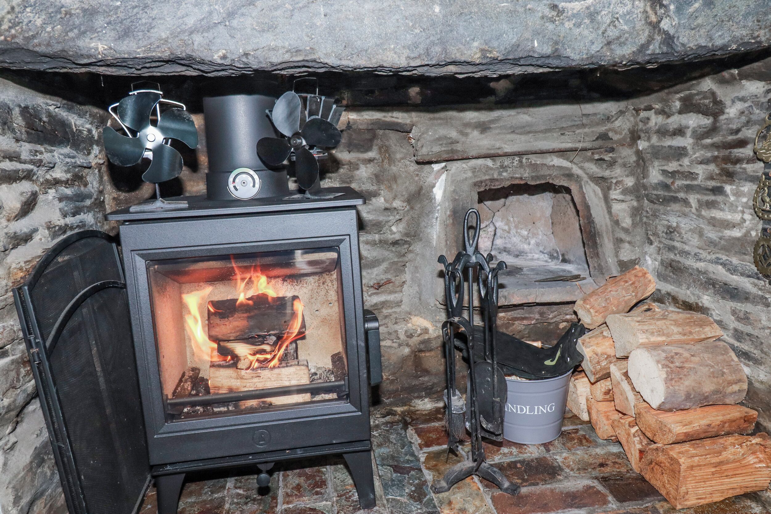 A wood-burning stove with a fire, a metal bucket labeled 'NORDLING,' and a stack of firewood.