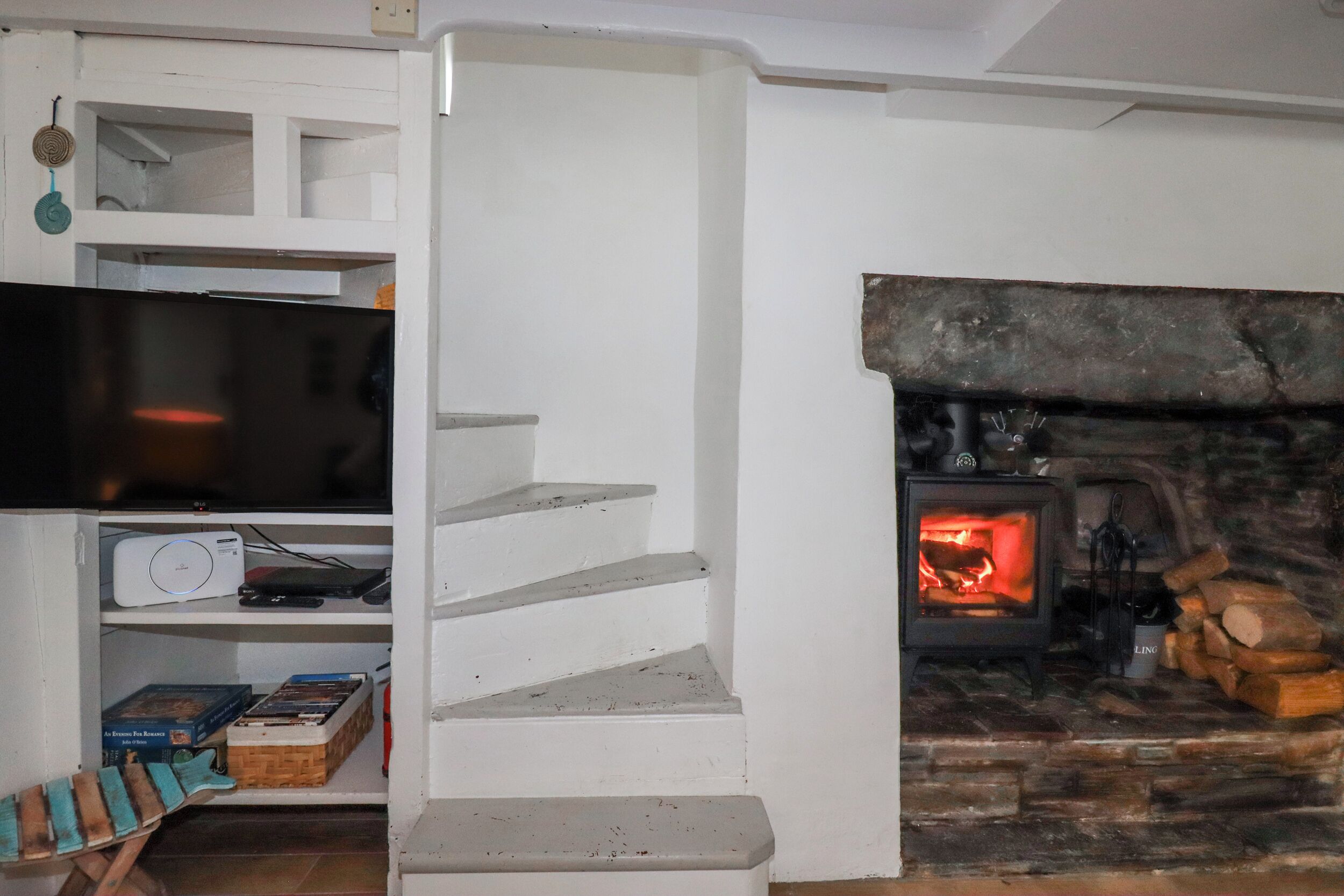 Una escalera conduce a una sala de estar con chimenea, televisor y una estantería con libros y un juego de mesa.
