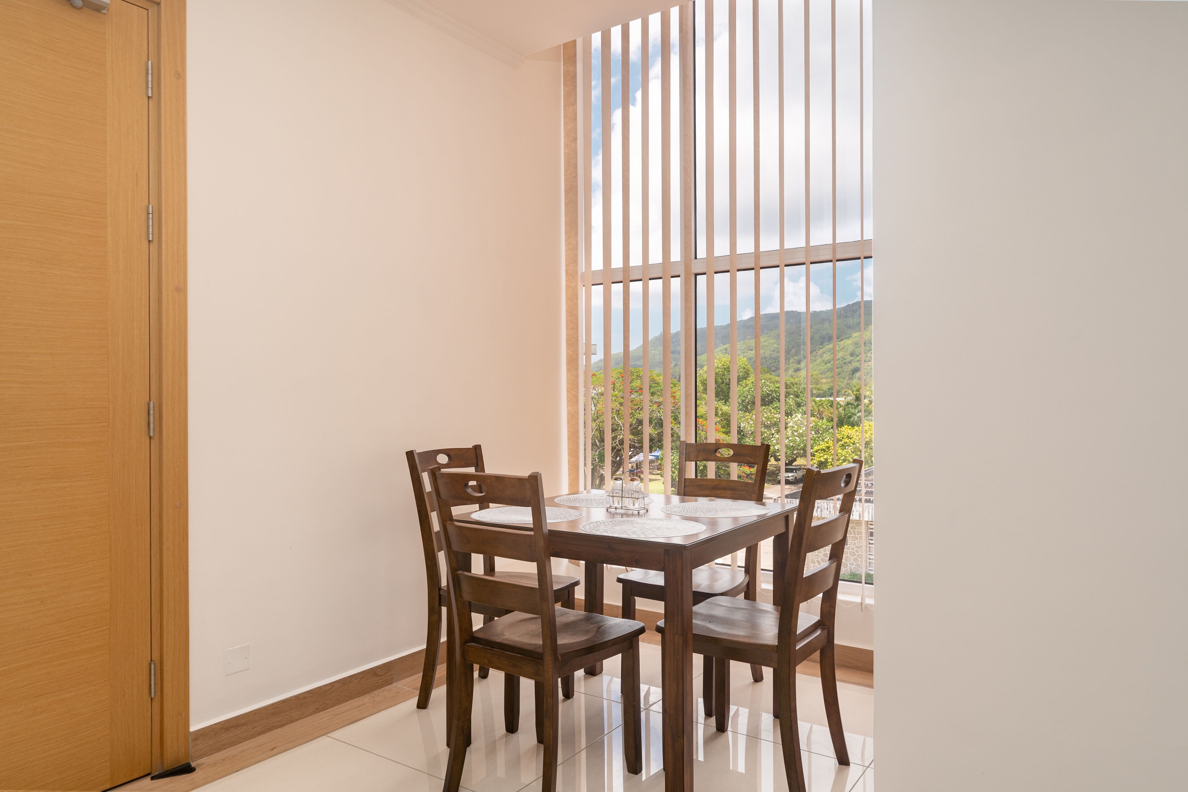 A dining area with a wooden table and chairs, a window with vertical blinds, and a view of greenery and mountains.