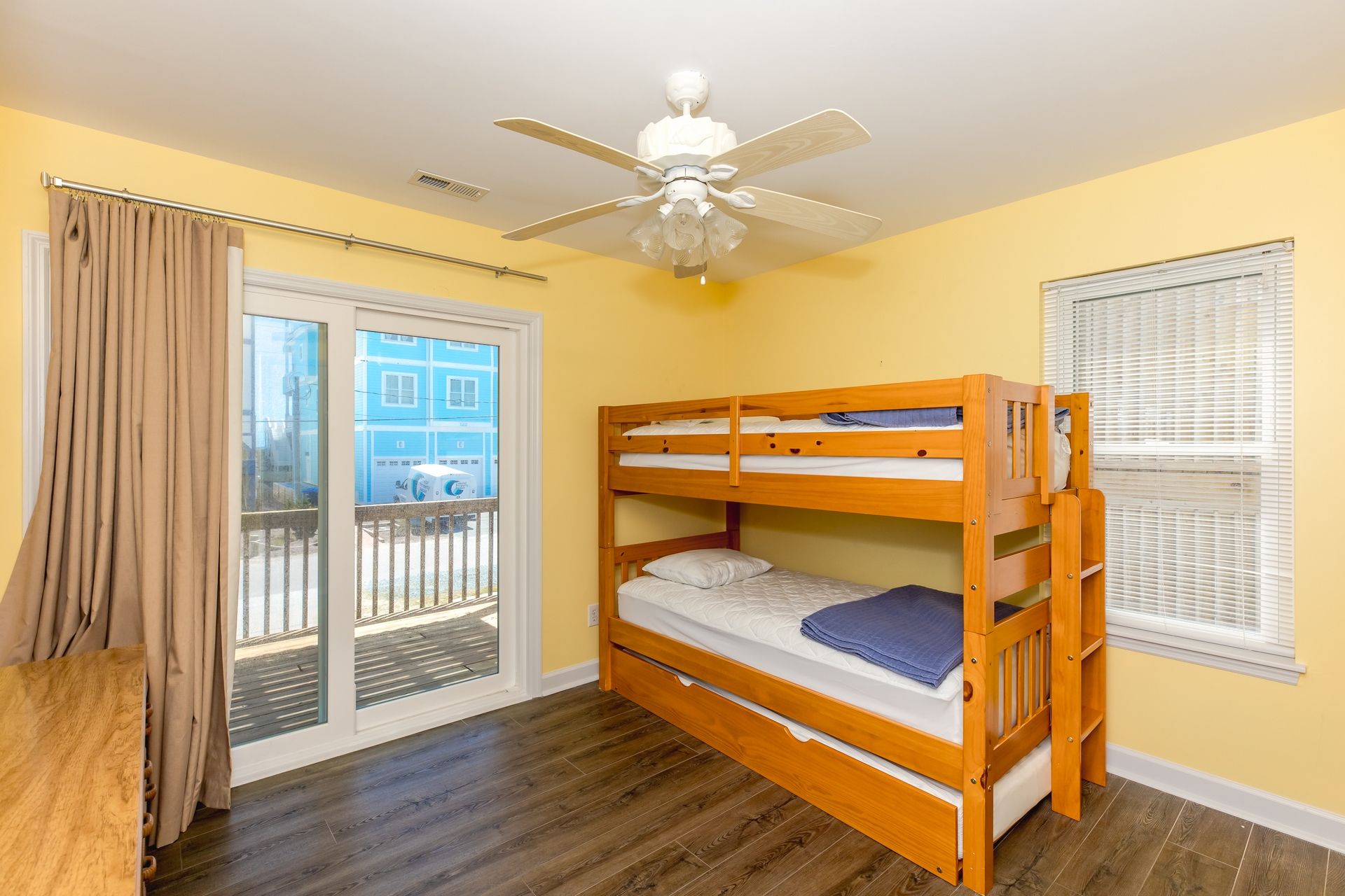 A room with a bunk bed, a ceiling fan, a window with blinds, and a view of buildings outside.