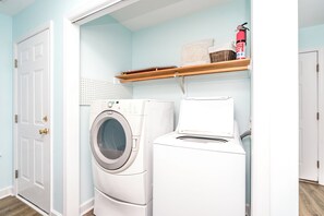 A laundry room with a front-loading washer and dryer, a shelf with a fire extinguisher, and a door with a gold handle. - No Regrets - 4 BR home in the heart of Surf City!  Dog Friendly! (Surf City)