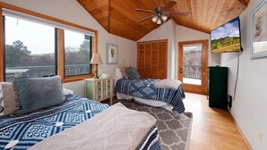 Une chambre avec deux lits, un ventilateur de plafond, des poutres apparentes, de grandes fenêtres et une armoire verte.