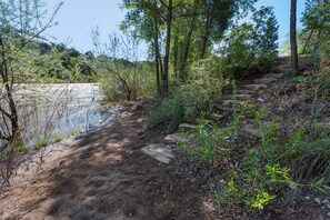 Un sentier de terre mène à une zone rocheuse, avec une rivière d’un côté et des arbres de l’autre.