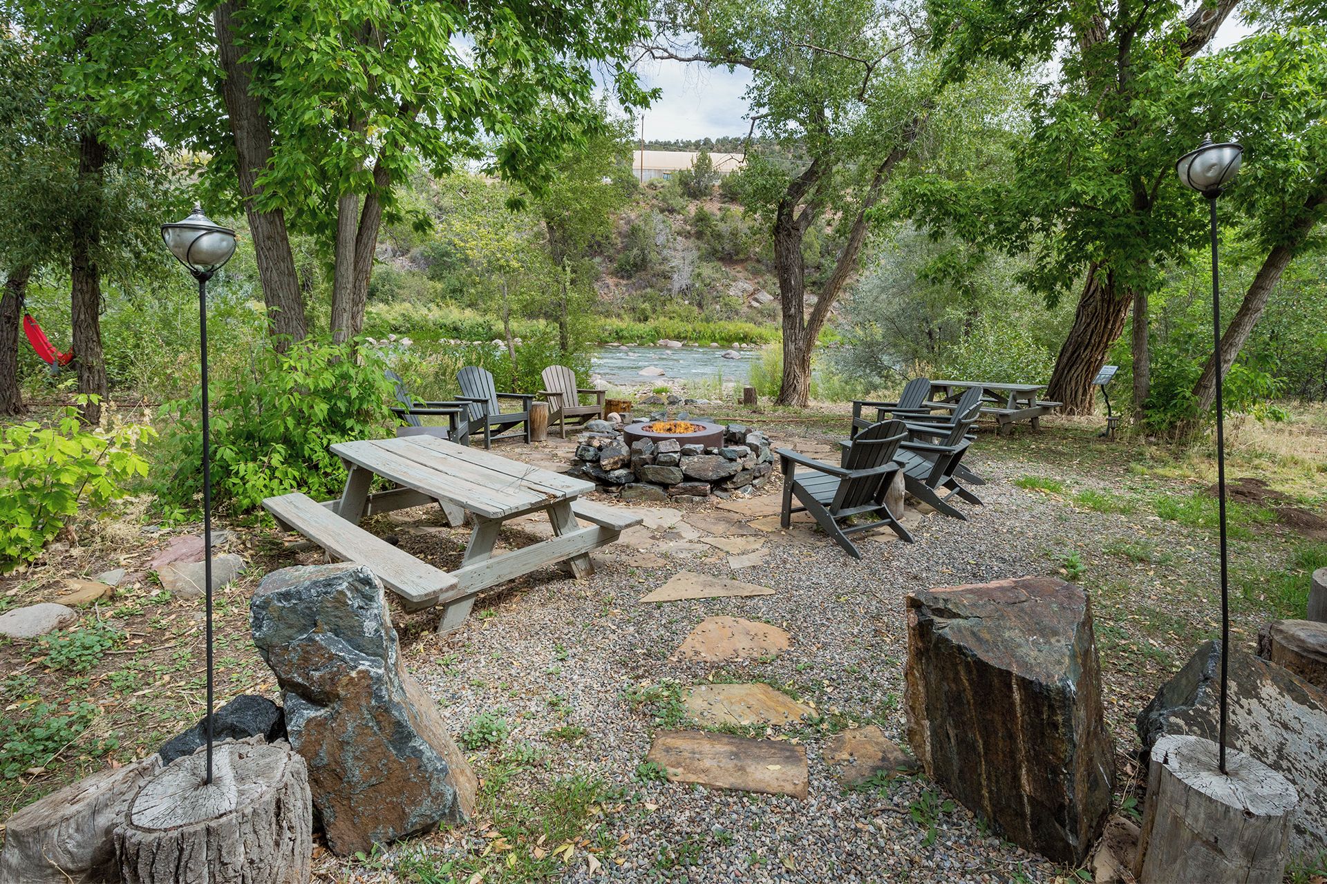 A rustic outdoor seating area with wooden benches and chairs, a stone fire pit, and a view of a river in the background.