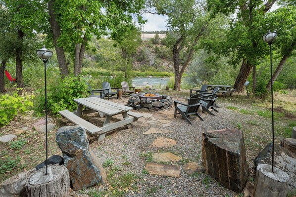 A rustic outdoor seating area with wooden benches and chairs, a stone fire pit, and a view of a river in the background.