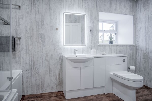 Uma casa de banho moderna com lavatório branco, sanita e um espelho grande com luz.