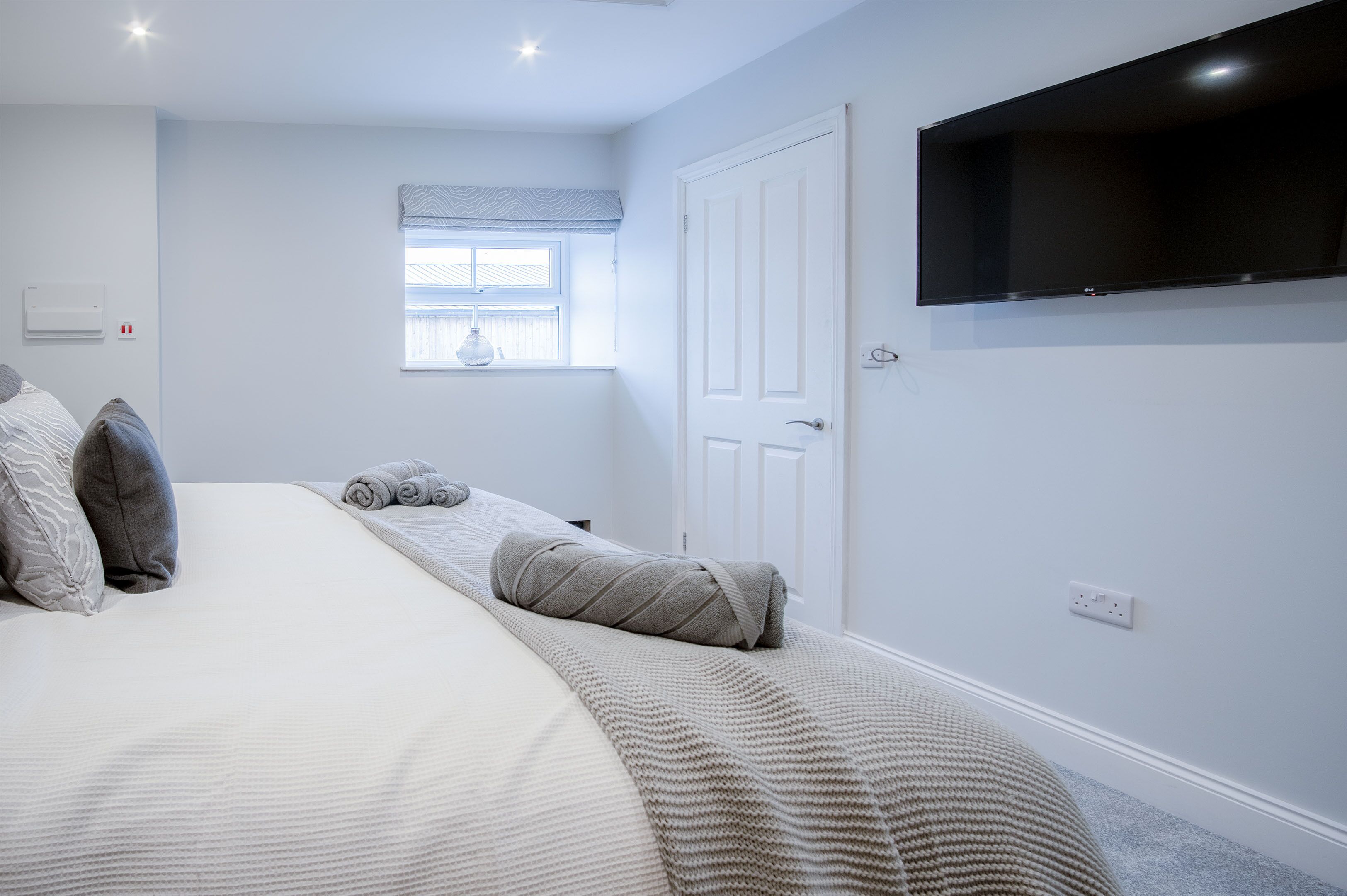 A bedroom with a bed, pillows, a window, a door, and a mounted TV.