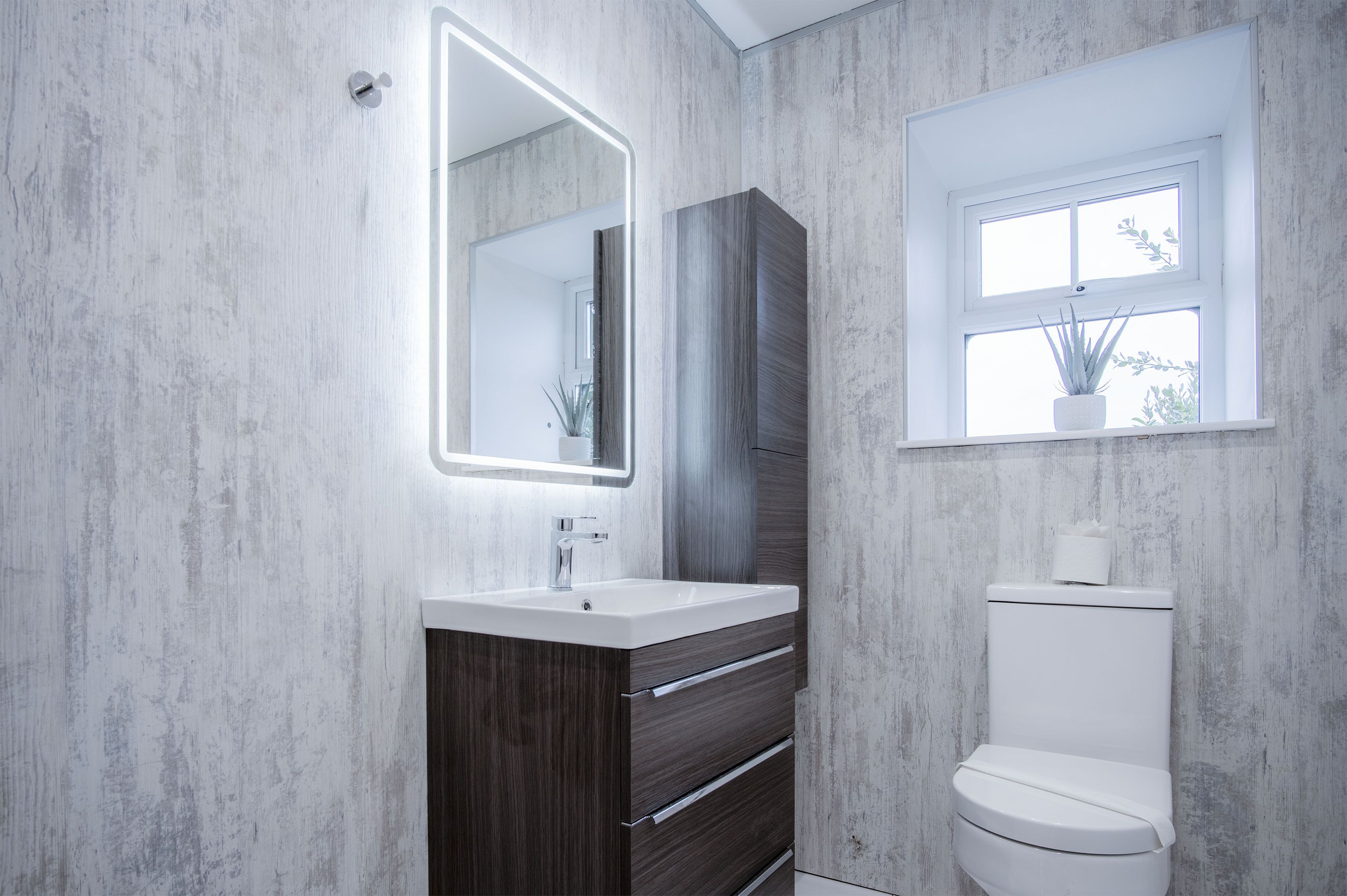 Une salle de bain moderne avec un lavabo blanc, un meuble-lavabo en bois foncé, un grand miroir doté d’un éclairage intégré, des toilettes blanches et une fenêtre agrémentée d’une plante en pot.