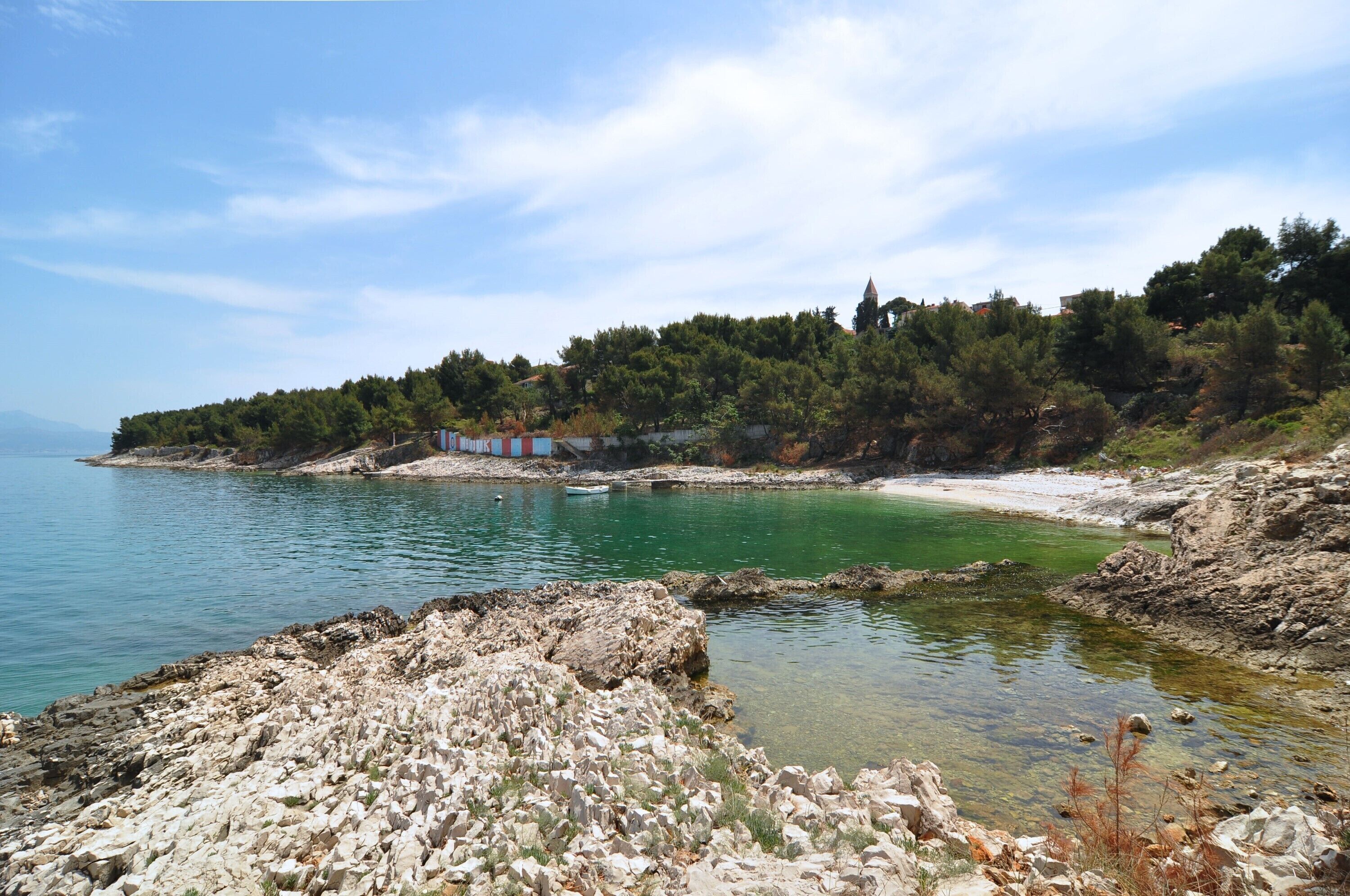 A coastal landscape with rocky terrain, clear water, and a forested hill in the background.