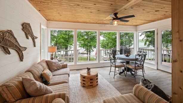 A cozy room with a wooden ceiling, large windows, a ceiling fan, a dining table with chairs, and a sofa.