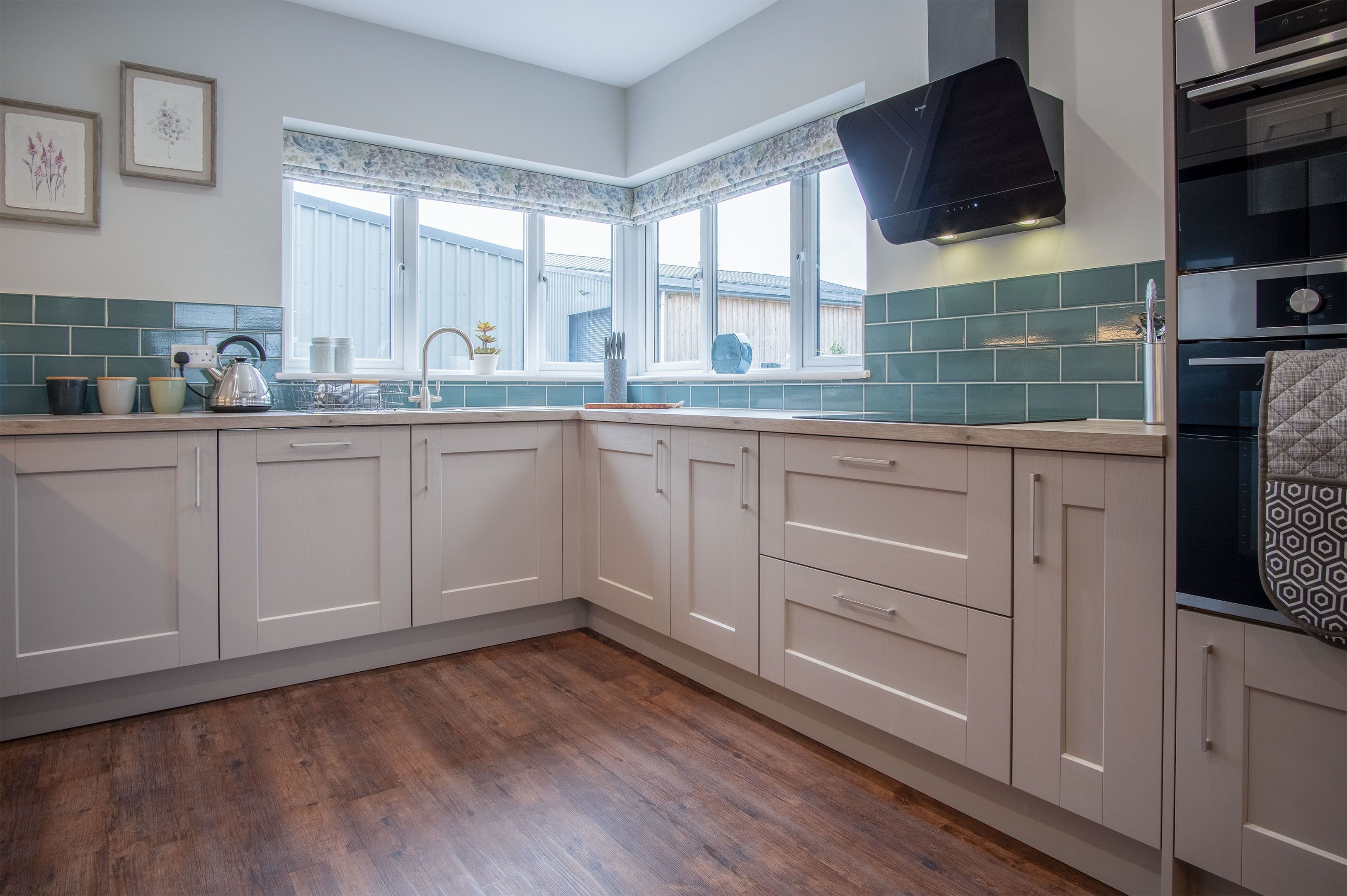 A kitchen with white cabinets, a window with a floral valance, a stainless steel range hood, and a tiled backsplash.