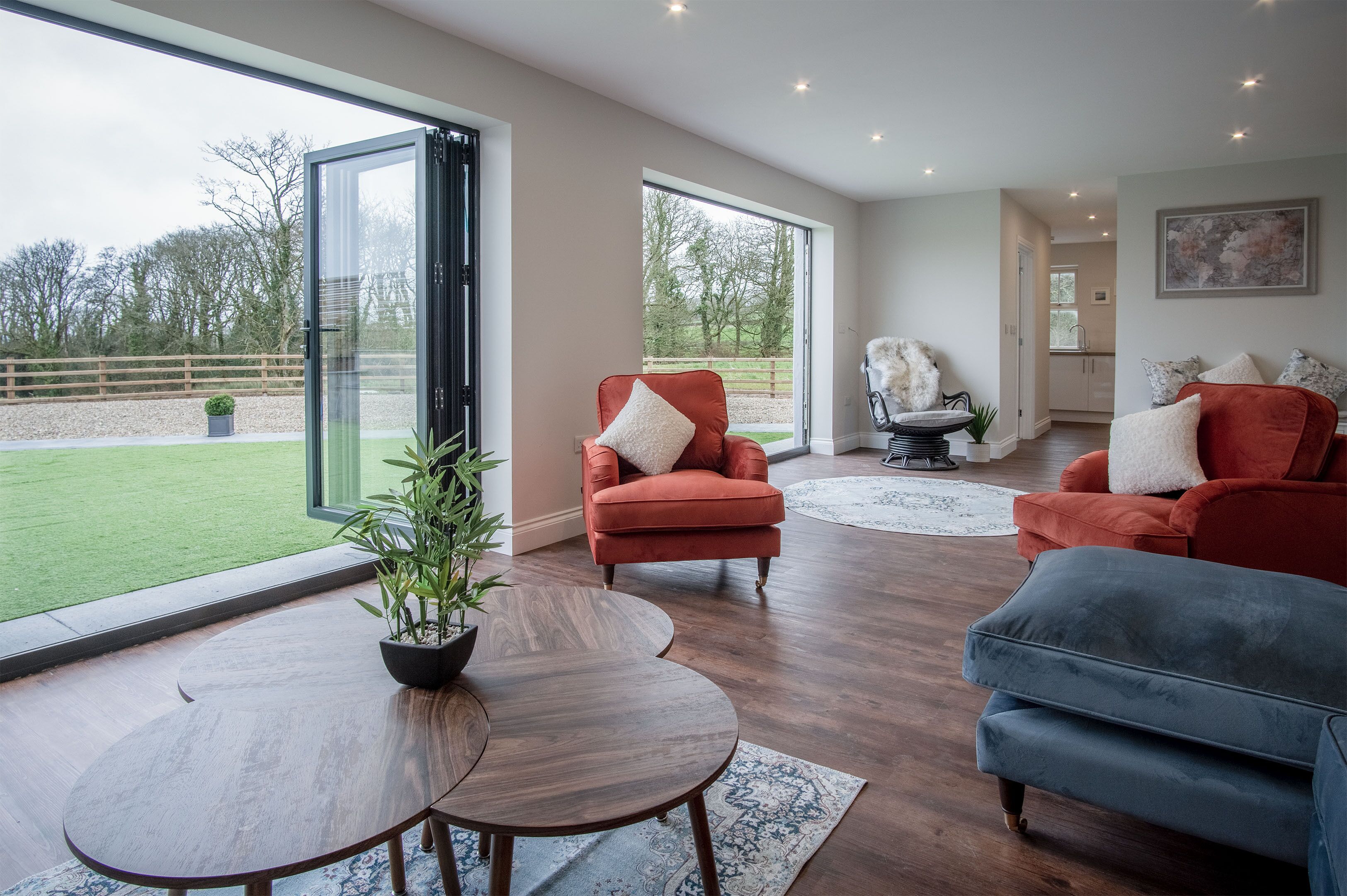 A spacious living room with a large sliding glass door leading to a green lawn, a wooden coffee table, and two sofas with cushions.