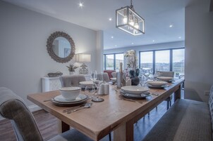 A dining area with a long wooden table set for four, a large mirror, and a view of the city through large windows.