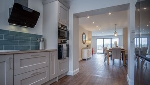 A modern kitchen with white cabinetry, a built-in oven, and a backsplash. There is a dining area with a table and chairs, and a large window providing natural light. - Kiln Park Farmhouse - 4 Bedroom Cottage - Narberth (Narberth)