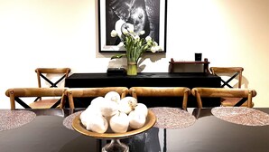 A dining table with a black tablecloth, surrounded by wooden chairs with woven seat cushions. A vase with white flowers is placed on the table, and a framed black and white photograph hangs on the wall behind. - Sarovar Holidays (Traralgon)