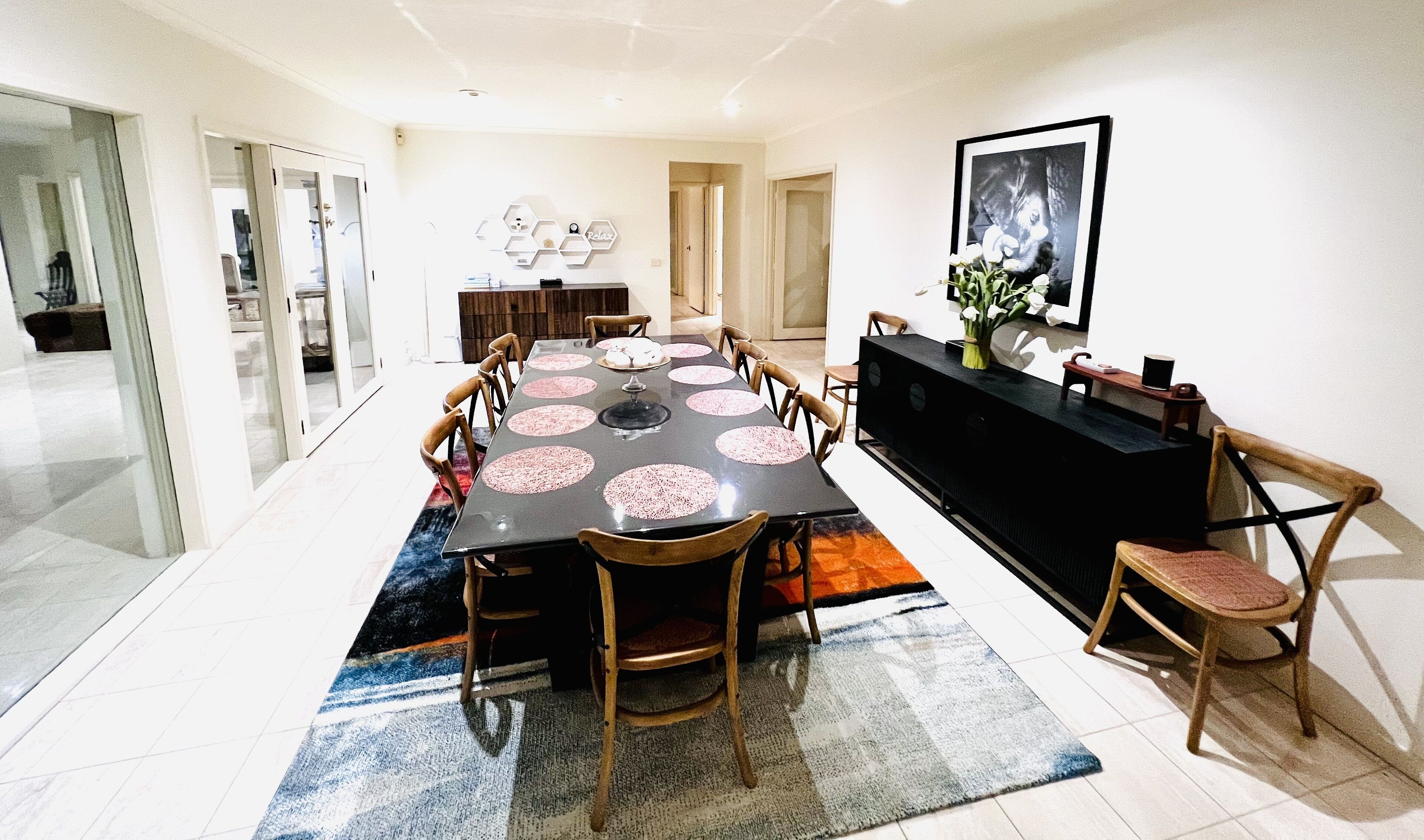 A modern dining area with a long table, multiple chairs, and a black console table with a vase of flowers.