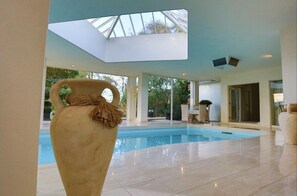Un vaste espace intérieur abritant une piscine, avec un plafond haut et une verrière, le tout entouré d’une architecture moderne et agrémenté d’un grand vase.