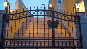 Un portail en fer forgé orné d’un blason, deux lampes fixées au mur et un escalier en pierre menant à l’entrée d’un bâtiment.
