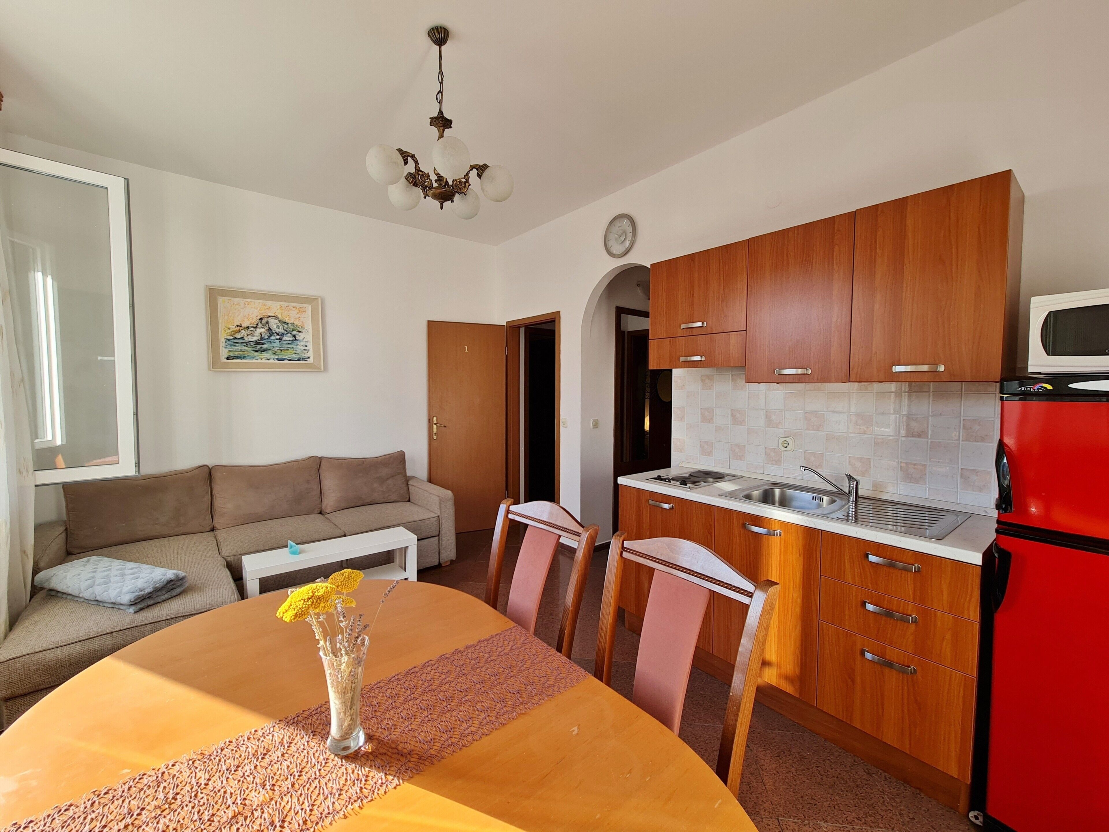 Un espacio habitable compacto con una mesa de comedor, sillas, un sofá, una zona de cocina con armarios de madera, un refrigerador rojo y una lámpara en el techo.