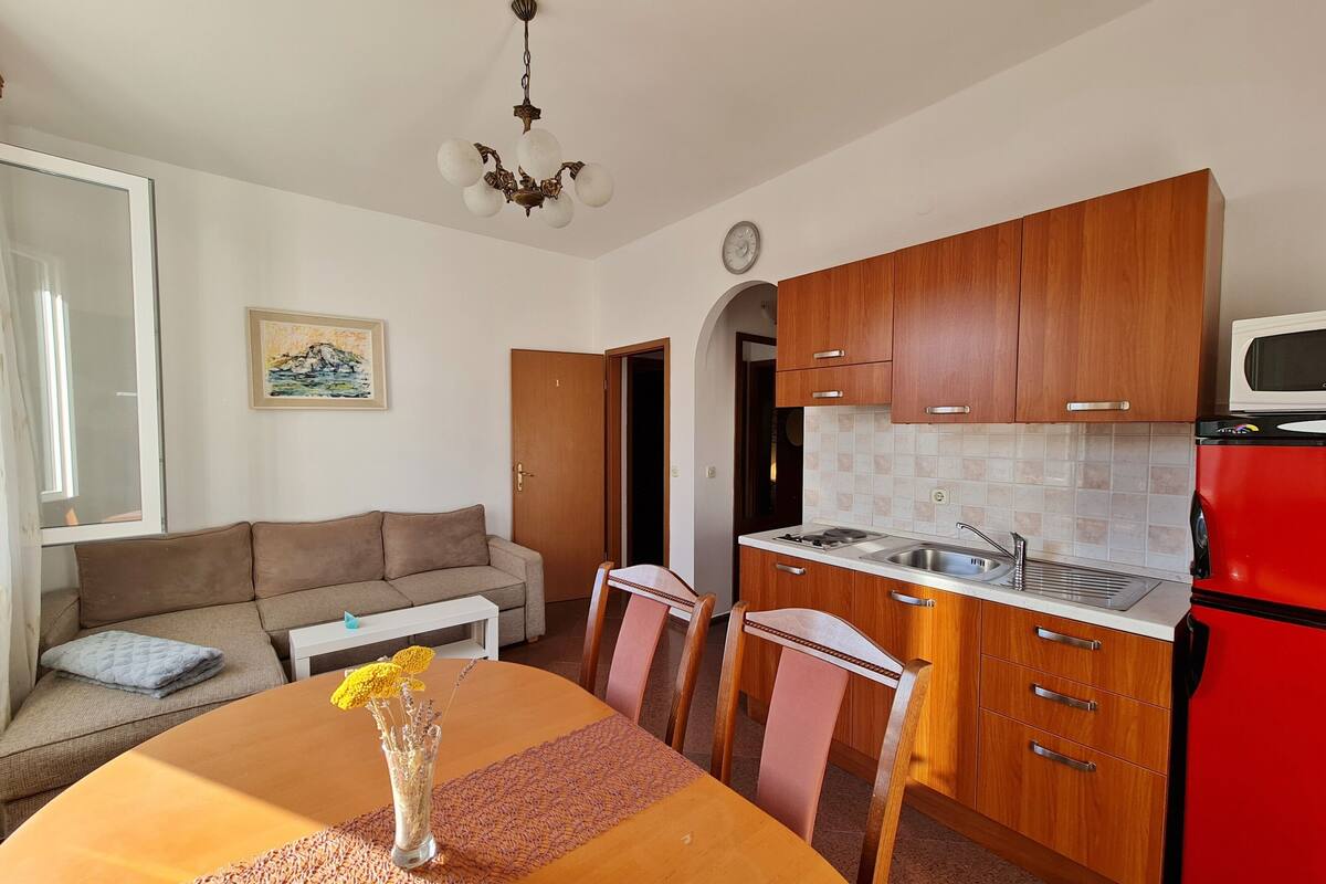 A compact living space with a dining table, chairs, a sofa, a kitchen area with wooden cabinets, a red refrigerator, and a ceiling light fixture.