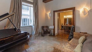 A spacious living room with a grand piano, a comfortable sofa, and a dining area visible through an open doorway.