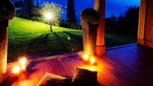A room with a wooden floor, two cushions, and a view of a well-lit garden at night.