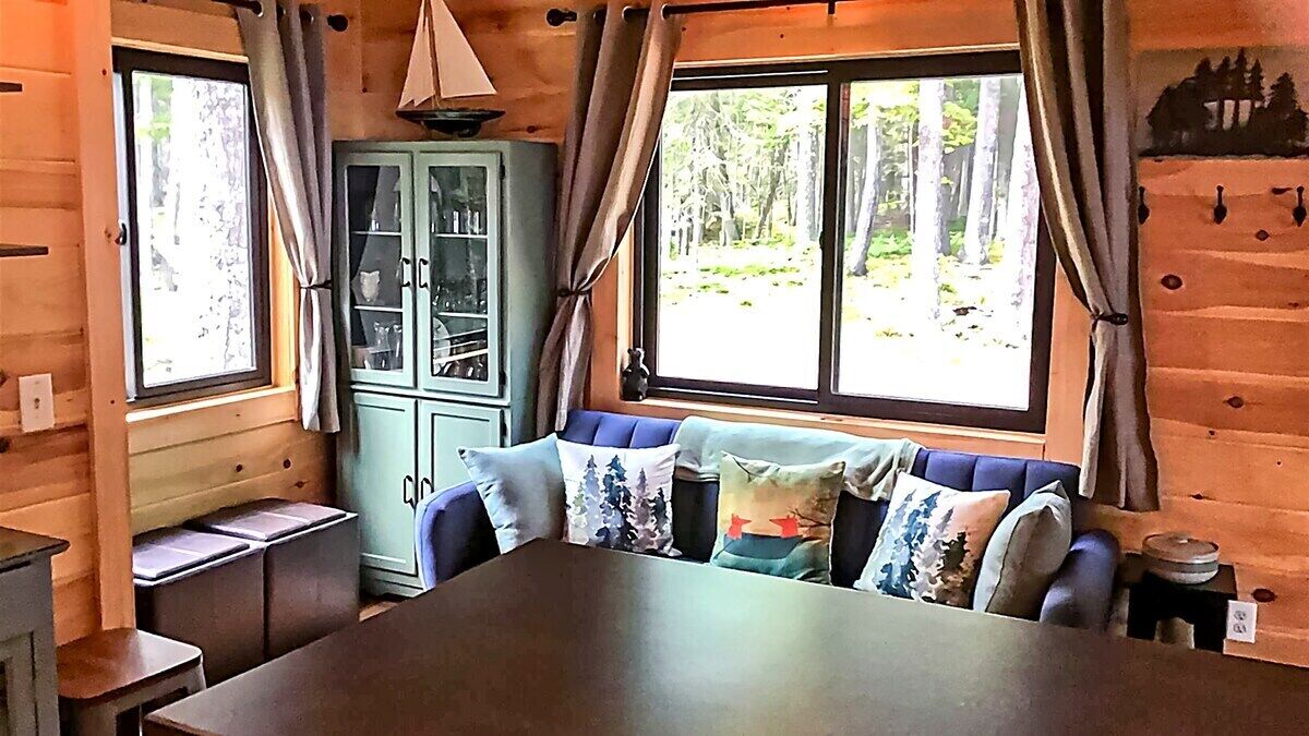 Ein Innenraum einer Holzhütte mit einem Esstisch, einer blauen Sitzbank, einem Schrank und Fenstern mit Vorhängen.