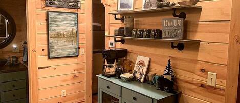 Un interior de madera con un cuadro enmarcado, estantes con objetos decorativos, una cafetera y un mueble pequeño.