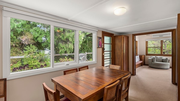 A dining area with a wooden table and chairs, large windows with a view of greenery, a wooden door, and a sofa in the adjacent room.