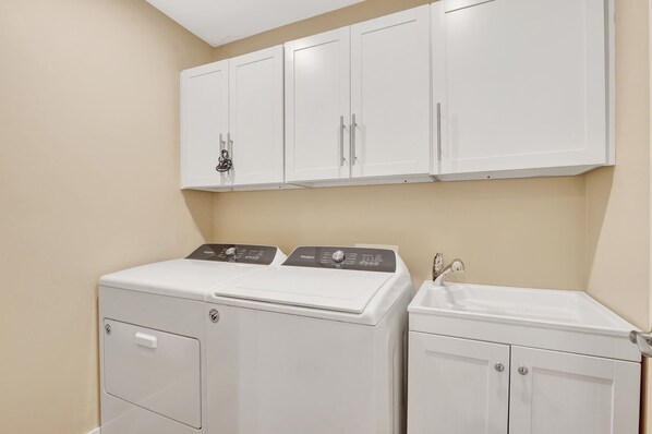 A laundry room with a front-loading washer and dryer, a sink, and white cabinets above. - Subtropical Oasis -Naples, Fl Retreat (Naples)