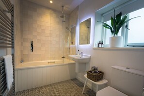 A modern bathroom with a bathtub, shower, sink, and toilet. There is a window with a plant and toiletries on the windowsill. - Luxury bolthole in secluded valley near coast (AXMINSTER)