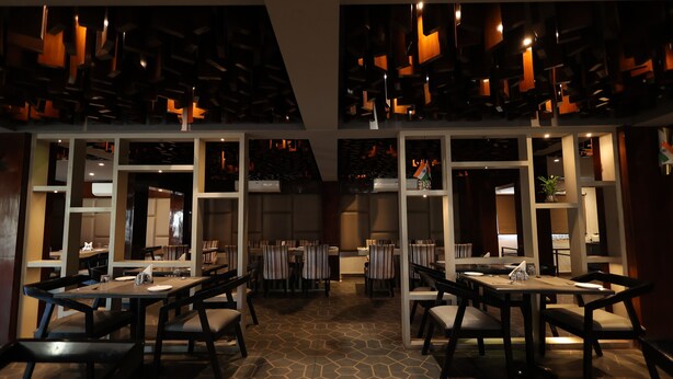 A modern restaurant with high ceilings, geometric ceiling design, and a central open space with tables and chairs. - THE LATITUDE LUXURY  (Haridwar)
