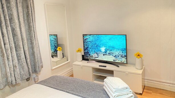 Quarto com cama, televisão sobre suporte branco, espelho e janela com cortinas.