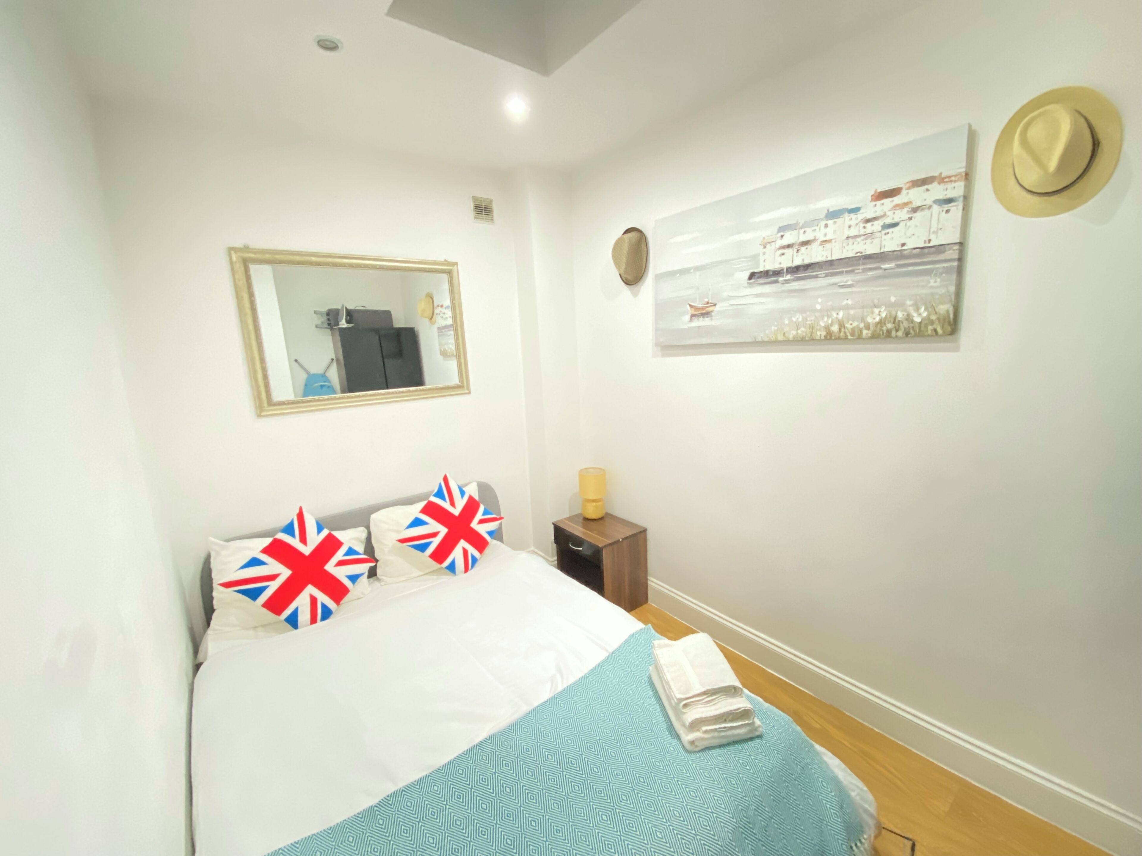 A neatly made bed with a blue and white patterned bedspread, two pillows with Union Jack designs, a small bedside table with a lamp, and a painting of a coastal scene on the wall.