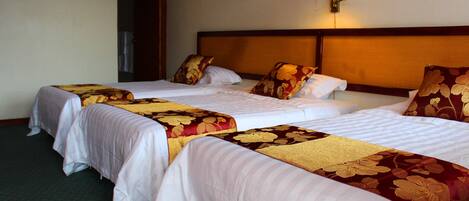 A hotel room with three beds, each with a different patterned bedspread. There is a wall-mounted lamp on the right side of the room.