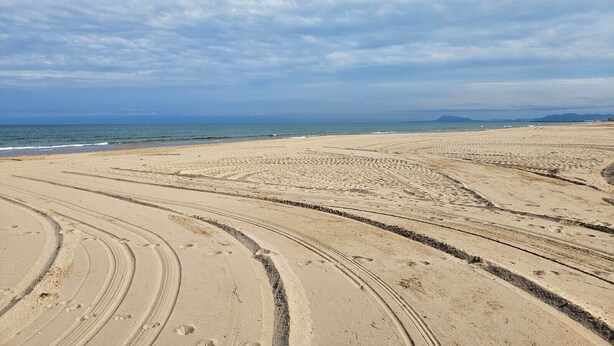 Ein weiter Sandstrand mit deutlich sichtbaren Radspuren und Fußabdrücken, der zum Meer führt.