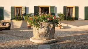 Un bac à fleurs en pierre, orné d’un motif décoratif, entouré d’une cour en gravier, devant un bâtiment aux volets verts et sur lequel est posé un banc.
