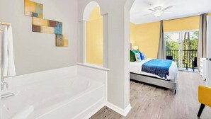 Un baño con bañera, ducha y ventana. Una habitación con cama, balcón y vistas a árboles.