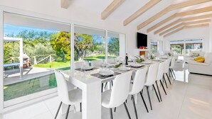 A modern dining area with a long table, white chairs, and large windows offering a view of a garden and outdoor seating. - Amazing Villa Chimera White View Pool (Arezzo)